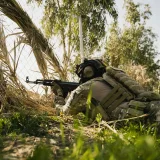 A soldier in tactical gear aims a rifle during an outdoor military exercise in a natural setting.
