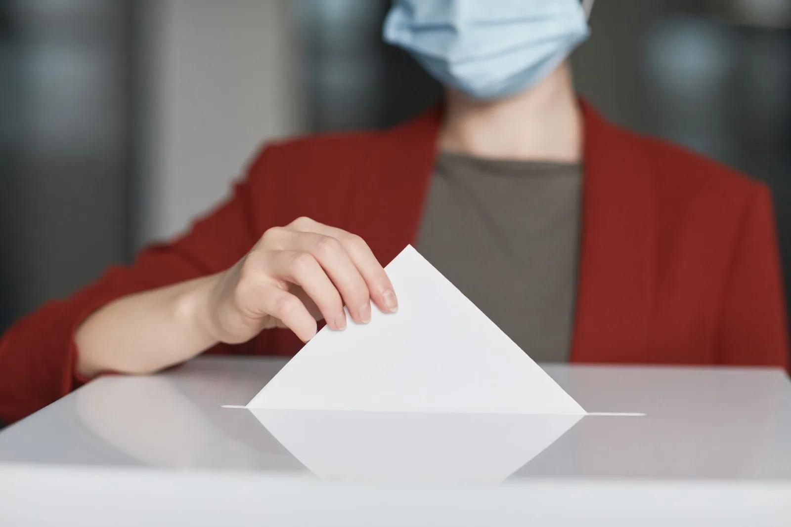 Persona con mascarilla depositando una papeleta en una urna para votar. Enfatiza el deber cívico.