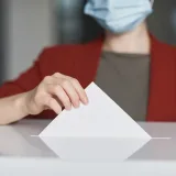 Persona con mascarilla depositando una papeleta en una urna para votar. Enfatiza el deber cívico.
