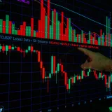 Primer plano de una mano señalando gráficos de criptomonedas en un monitor, como parte de un análisis de mercado.