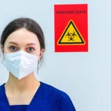 Healthcare professional in protective gear standing near a biohazard warning sign.