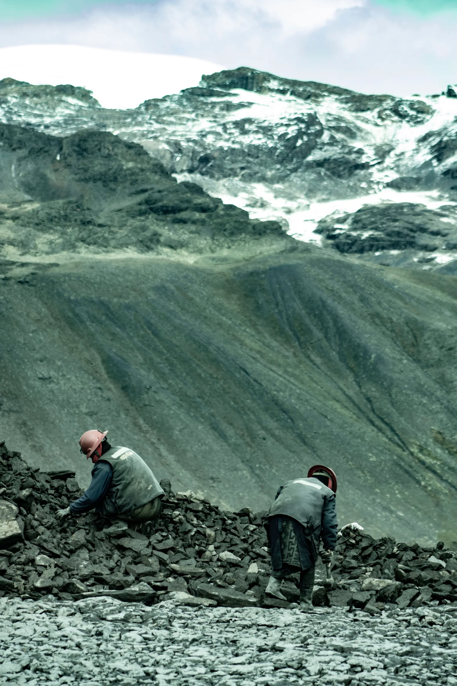 Dos mineros en Perú excavan entre montones de rocas en los Andes en busca de oro, bajo montañas nevadas.
