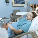 Mujer caucásica en cama de hospital recibiendo atención de un profesional de la salud.