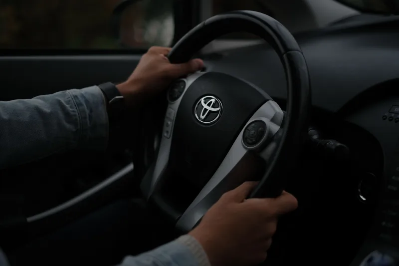 Manos sobre el volante de un Toyota, en primer plano dentro de un vehículo. Ambiente oscuro.