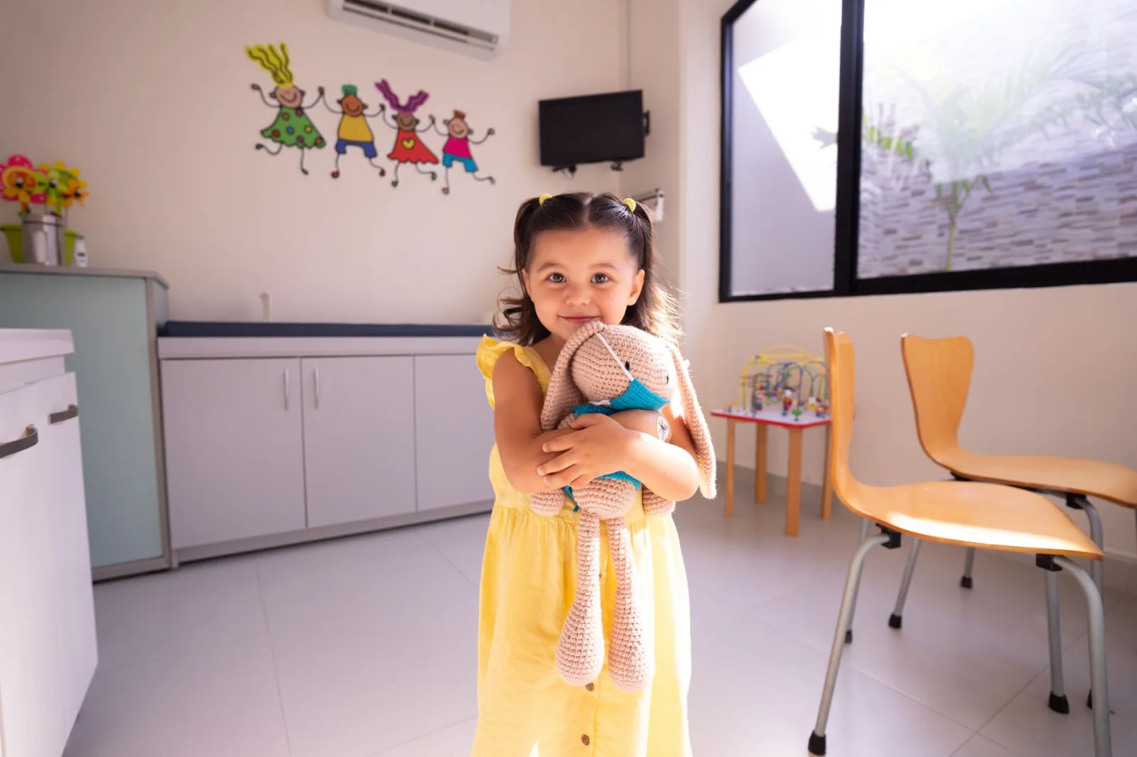 Una niña alegre con un vestido amarillo abraza un peluche en una habitación luminosa con una decoración divertida.