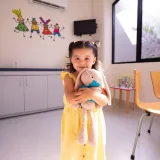 Una niña alegre con un vestido amarillo abraza un peluche en una habitación luminosa con una decoración divertida.