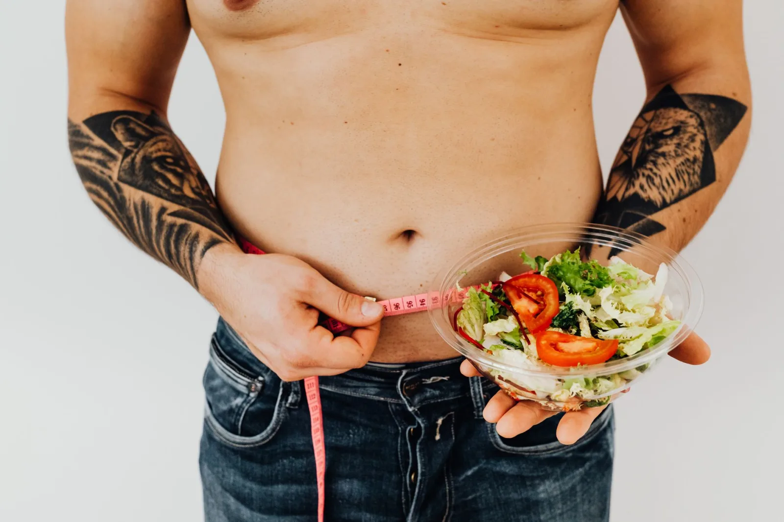Hombre tatuado midiéndose la cintura mientras sostiene una ensalada fresca, como símbolo de un estilo de vida saludable y el control del peso.