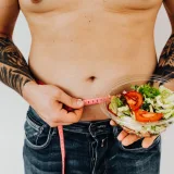 Hombre tatuado midiéndose la cintura mientras sostiene una ensalada fresca, como símbolo de un estilo de vida saludable y el control del peso.