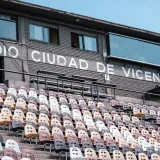 Empty stadium seats with a view of Estadio Ciudad de Vicente López sign, featuring filming equipment and operators.
