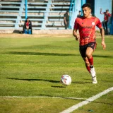 A young soccer player in red uniform dribbles the ball during a sunny match in Mendoza, Argentina.