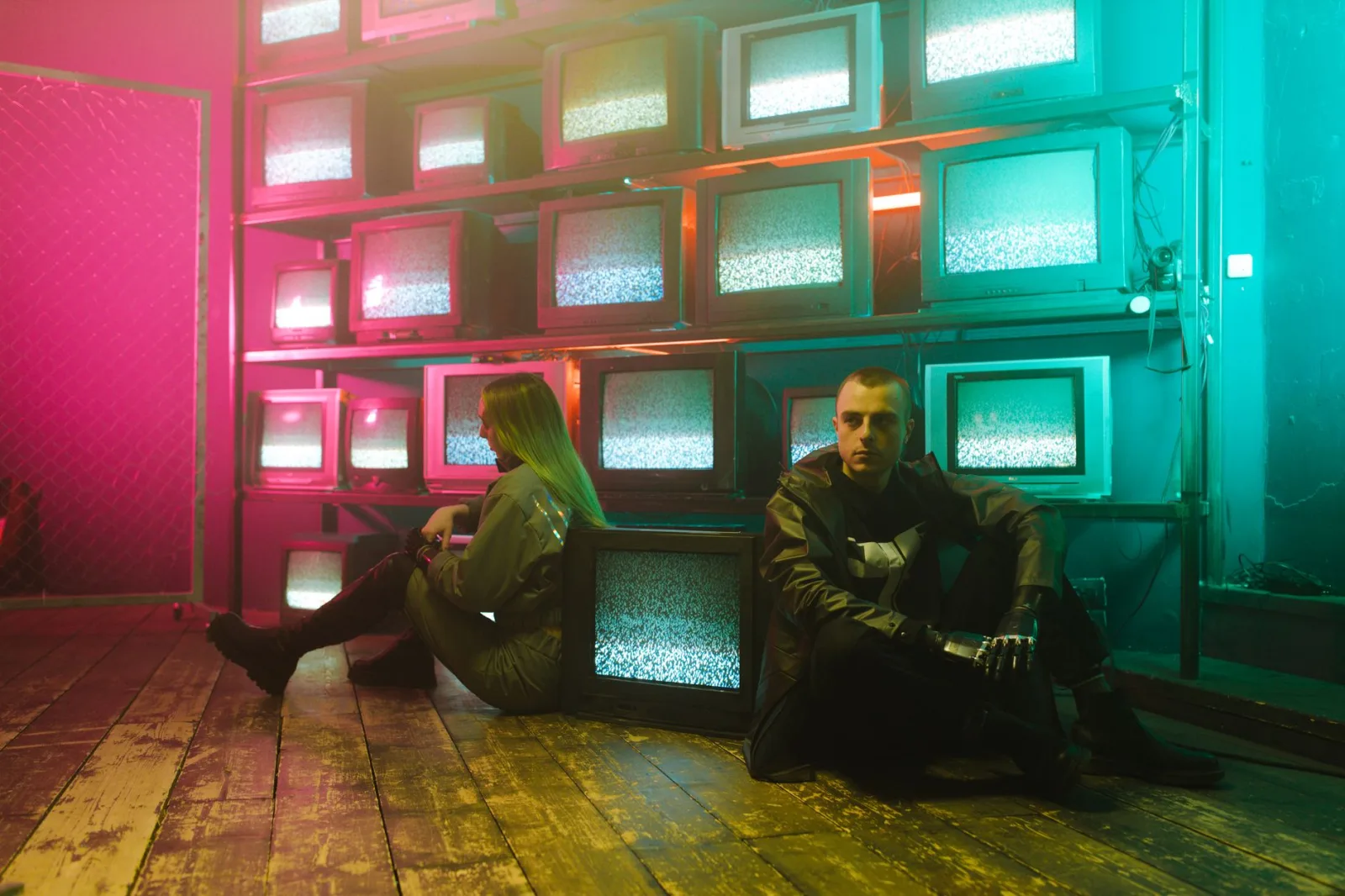 Dos individuos en un entorno cyberpunk con iluminación de neón y pantallas de televisión apiladas.