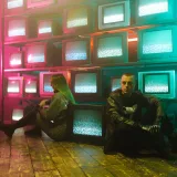 Dos individuos en un entorno cyberpunk con iluminación de neón y pantallas de televisión apiladas.
