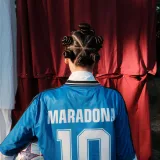 Vista trasera de una persona con una camiseta de Maradona y un peinado original, sosteniendo un balón de fútbol.