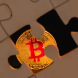 Una moneda de bitcoin parcialmente oculta bajo una pieza de rompecabezas sobre una superficie de madera, lo que simboliza la complejidad de la inversión digital.