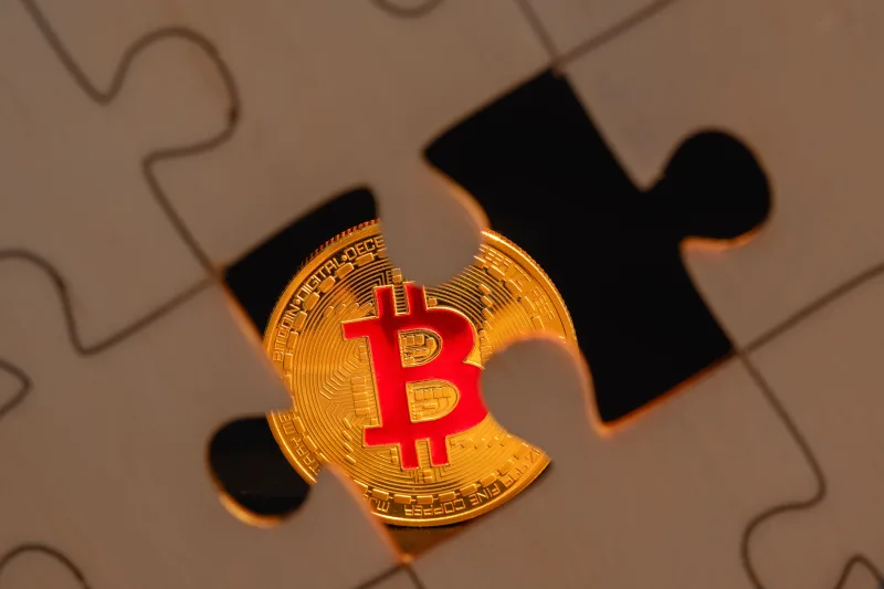 Una moneda de bitcoin parcialmente oculta bajo una pieza de rompecabezas sobre una superficie de madera, lo que simboliza la complejidad de la inversión digital.