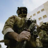 Un soldado con equipo táctico sostiene un rifle al aire libre, junto a unos edificios.