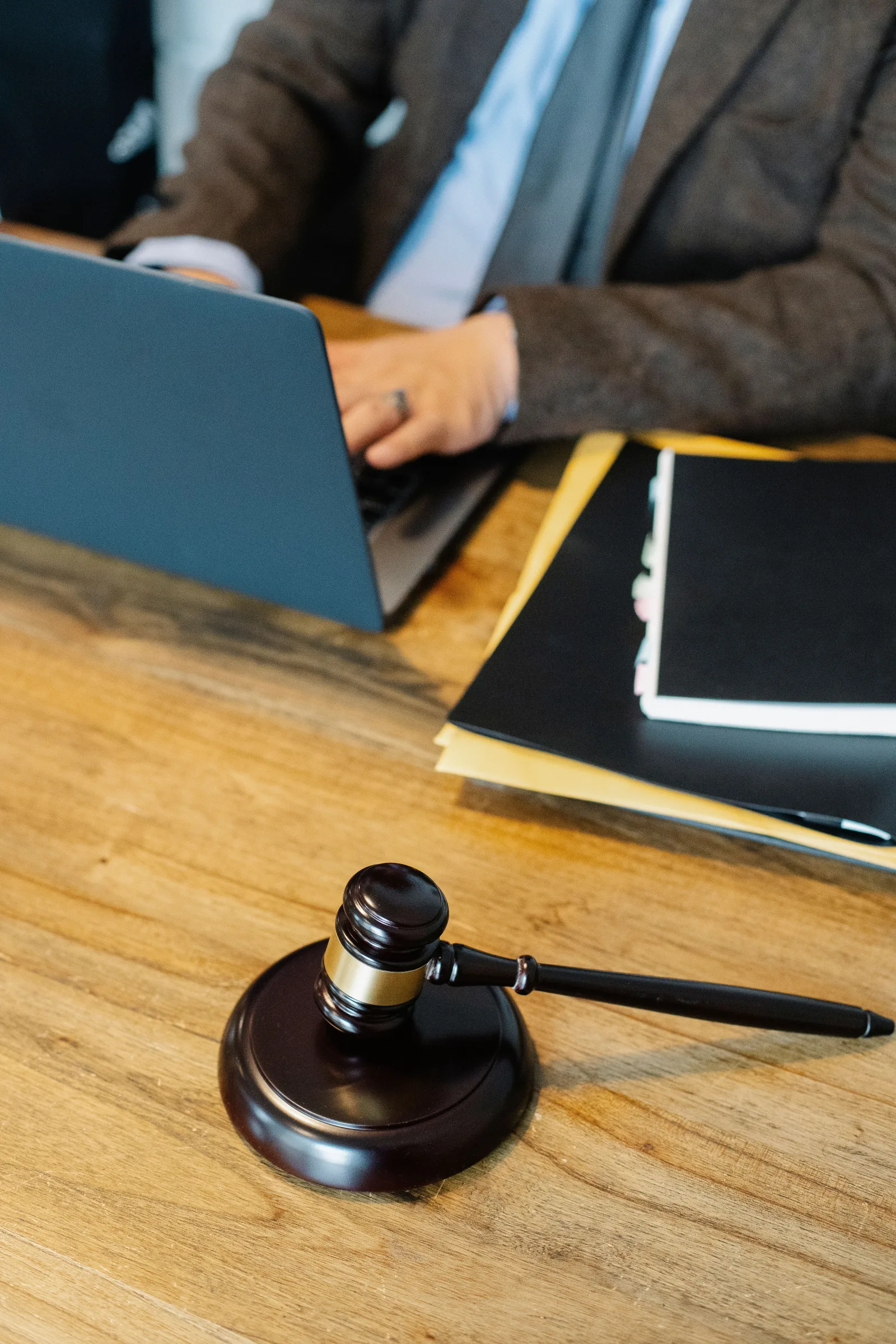 Vista cenital de un abogado anónimo vestido formalmente, escribiendo en una laptop mientras está sentado a una mesa de madera con una pila de documentos y un mazo judicial.