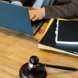 Vista cenital de un abogado anónimo vestido formalmente, escribiendo en una laptop mientras está sentado a una mesa de madera con una pila de documentos y un mazo judicial.