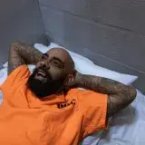 Hombre tatuado con traje naranja sonriendo mientras yace en una celda de prisión.