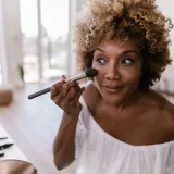 Woman with curly hair applying makeup inside a well-lit room, creating a natural look.