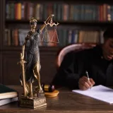 A focused judge writing on documents beside a Lady Justice statue in an office.