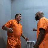Two male prisoners in orange jumpsuits talking inside a prison cell.