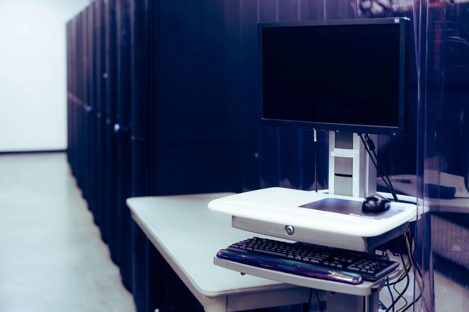 Computadora moderna con pantalla negra colocada sobre un soporte cerca de una fila de racks de servidores de acero en un centro de datos.