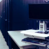 Computadora moderna con pantalla negra colocada sobre un soporte cerca de una fila de racks de servidores de acero en un centro de datos.