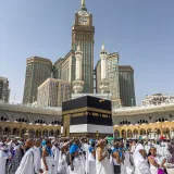 A large group of pilgrims gather at the Kaaba in Mecca, Saudi Arabia, during the Hajj pilgrimage under a clear blue sky.