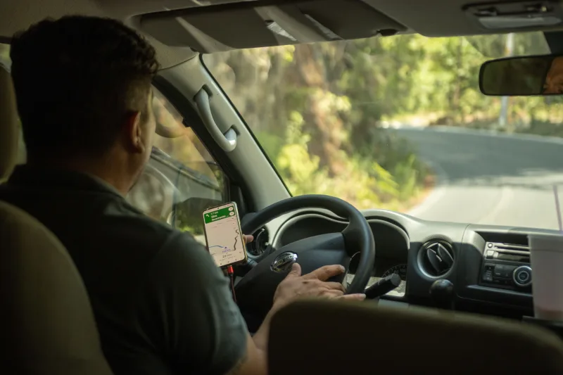 Conductor que utiliza una aplicación de GPS en su teléfono móvil para navegar por una carretera sinuosa rodeada de naturaleza.