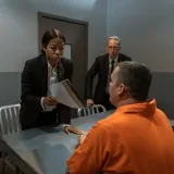Una tensa escena de interrogatorio en una sala de prisión con oficiales y un prisionero.