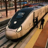Un tren AVE de alta velocidad detenido en una plataforma urbana, con pasajeros subiendo a bordo.