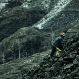 A solitary worker climbs a rocky slope in a barren quarry, wearing safety gear.