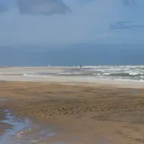 Escena tranquila de playa con olas suaves y personas caminando a lo lejos bajo un cielo azul brillante.