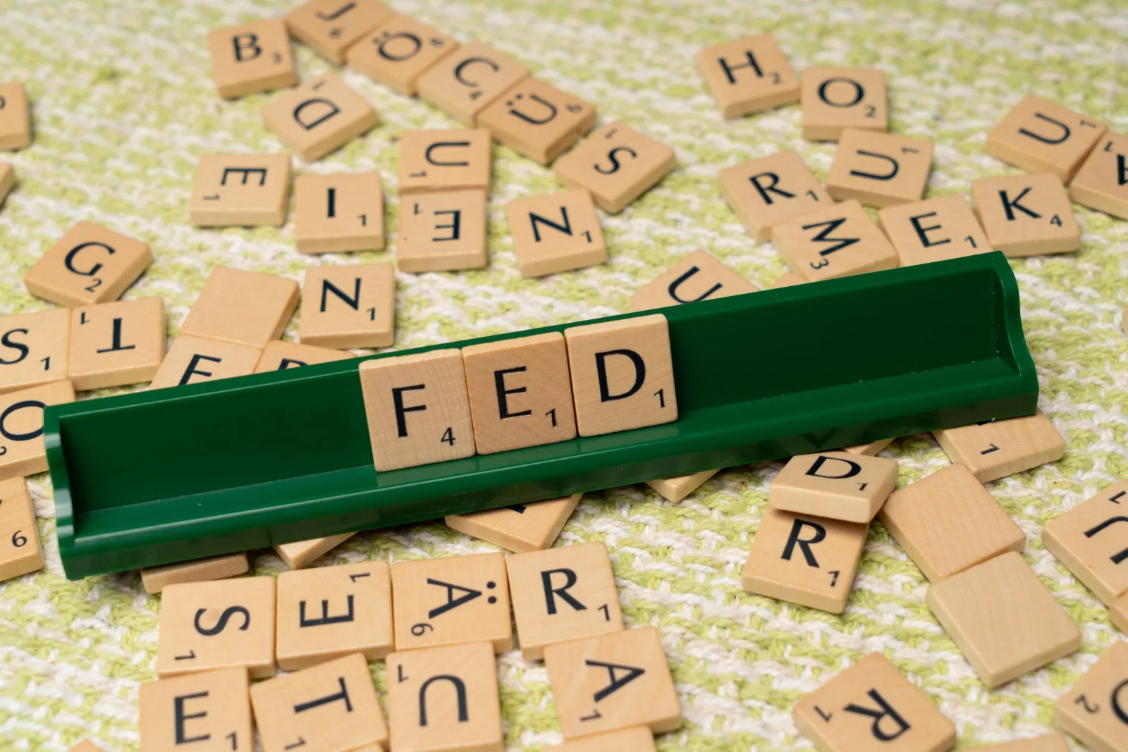 Fichas de Scrabble que forman la palabra «FED» en un atril verde, rodeadas de fichas dispersas sobre una superficie texturizada.