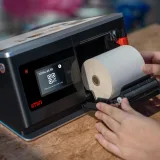 Manos colocando un rollo térmico en un dispositivo de punto de venta con pantalla táctil.
