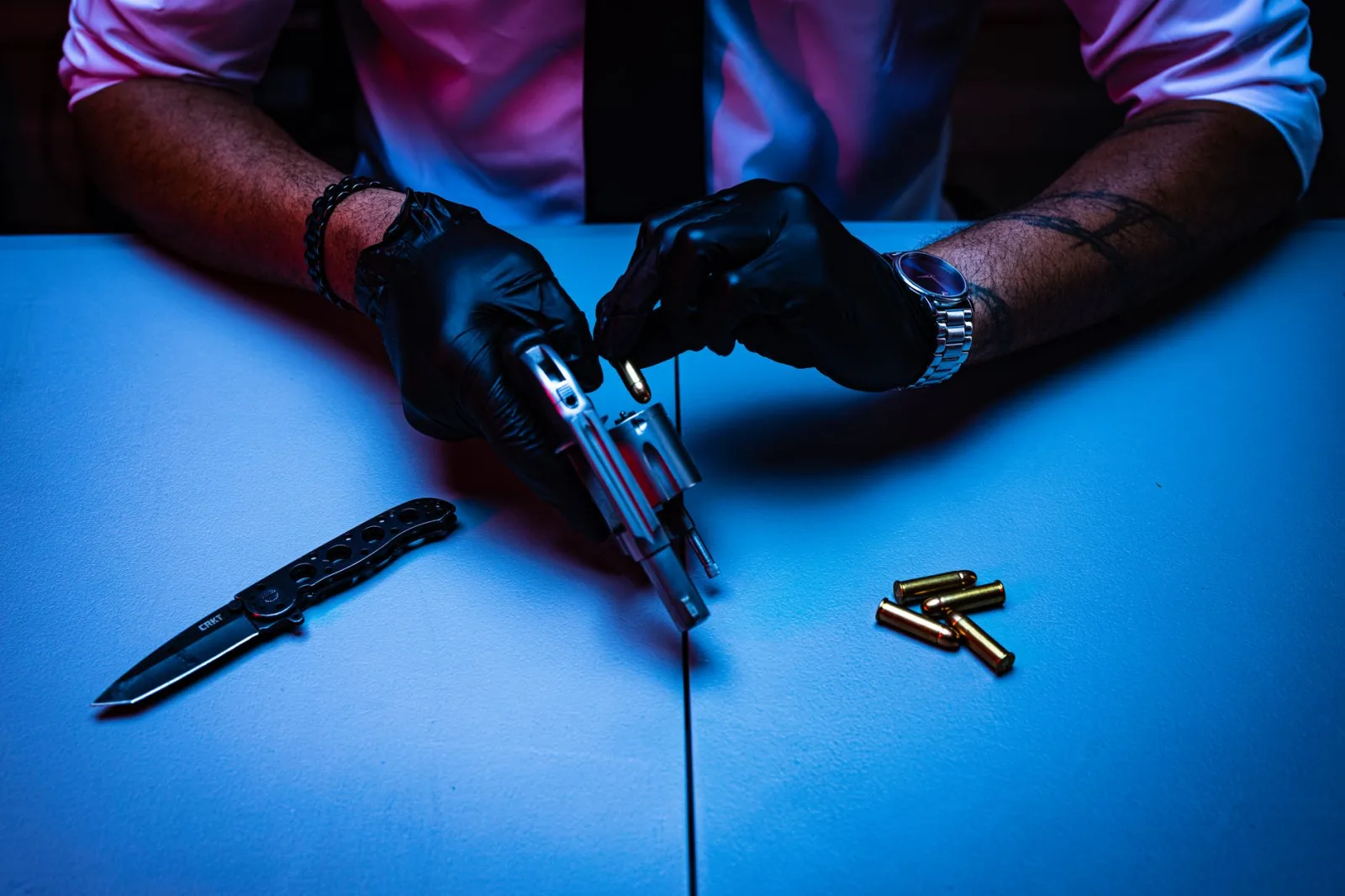 Hombre con guantes manipulando un revólver y un cuchillo sobre una mesa, con balas cerca.