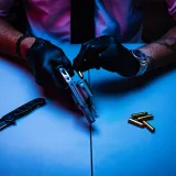 Hombre con guantes manipulando un revólver y un cuchillo sobre una mesa, con balas cerca.