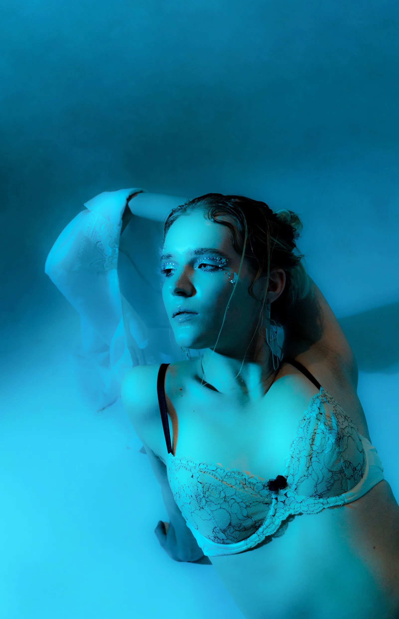 Mujer seductora en ángulo alto, con sujetador blanco, sentada en el suelo de una habitación oscura iluminada con luces azules, mirando hacia otro lado con sensualidad