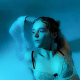 Mujer seductora en ángulo alto, con sujetador blanco, sentada en el suelo de una habitación oscura iluminada con luces azules, mirando hacia otro lado con sensualidad