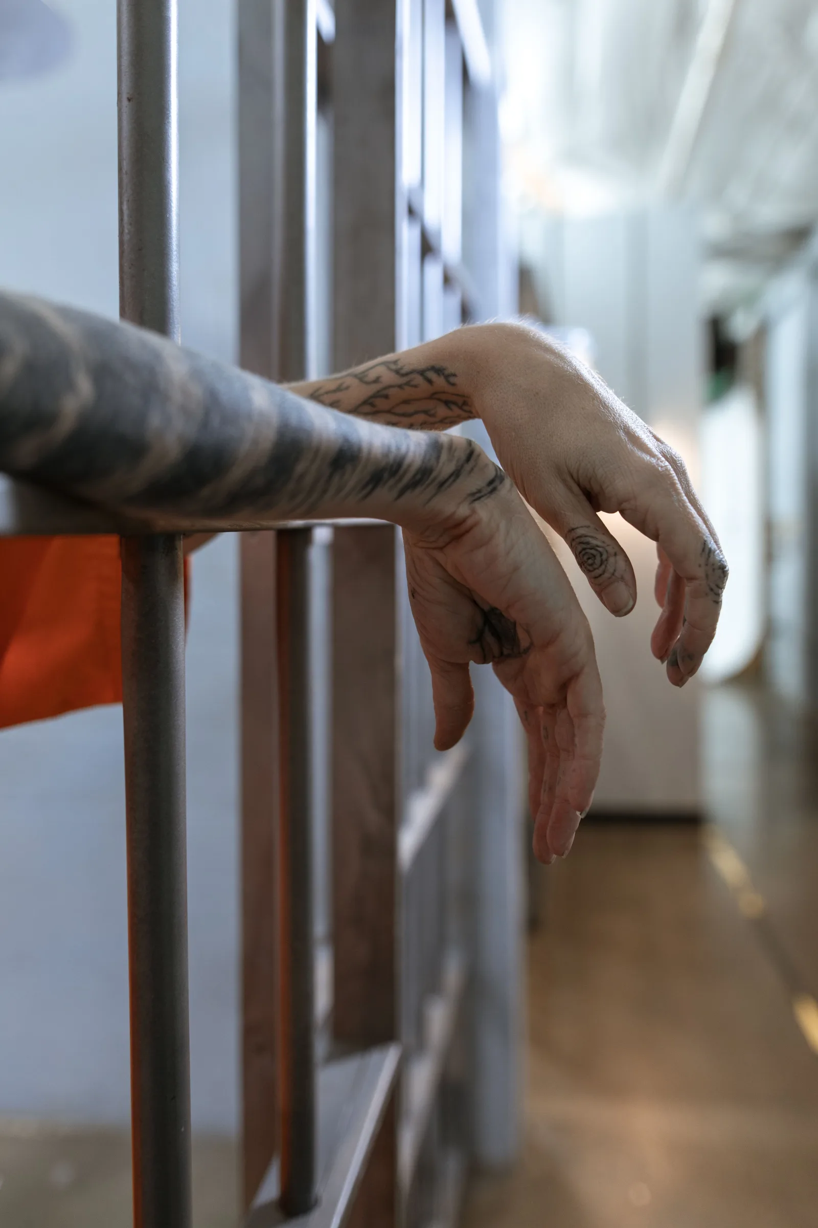 Primer plano de unas manos tatuadas colgando detrás de los barrotes de una celda, evocando el confinamiento.
