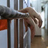 Primer plano de unas manos tatuadas colgando detrás de los barrotes de una celda, evocando el confinamiento.