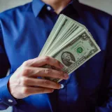 Primer plano de una persona con camisa azul sosteniendo varios billetes de dólares estadounidenses.