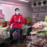 Hombre asiático con chaqueta roja y mascarilla trabajando en una tienda con carritos de la compra en Wuhan, China.