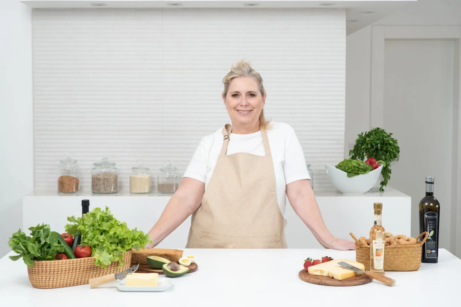 Una mujer adulta con delantal está de pie frente a la encimera de la cocina, con productos frescos y queso, lista para cocinar.
