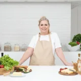 Una mujer adulta con delantal está de pie frente a la encimera de la cocina, con productos frescos y queso, lista para cocinar.