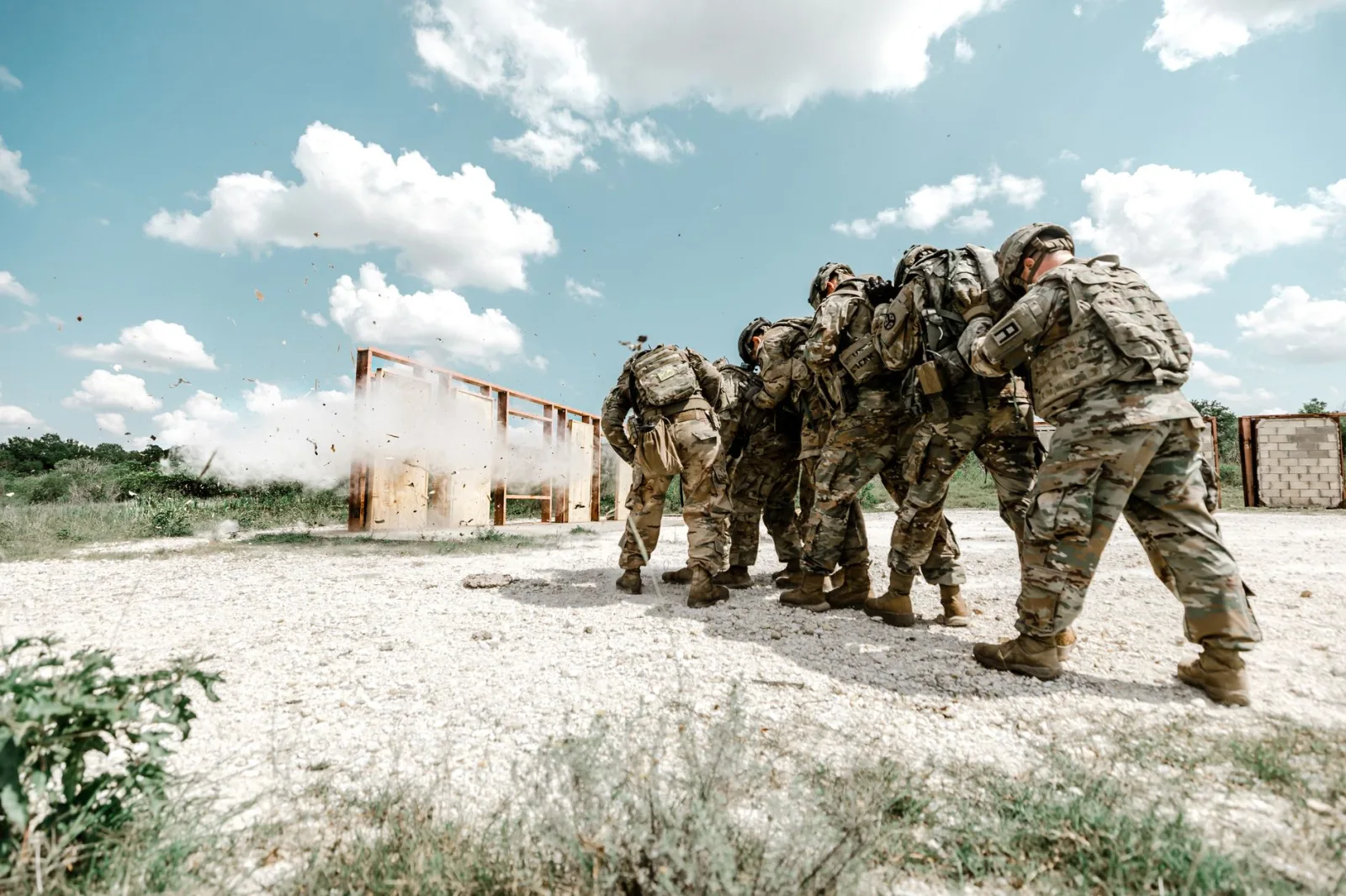 Grupo de soldados con uniformes militares realizando un ejercicio de entrenamiento al aire libre con simulaciones de explosiones.