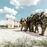 Grupo de soldados con uniformes militares realizando un ejercicio de entrenamiento al aire libre con simulaciones de explosiones.