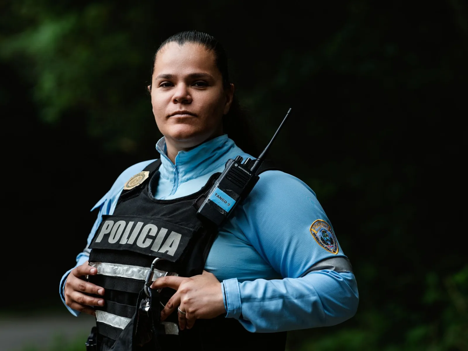 Female police officer in uniform standing outdoors with a confident expression and radio handset.