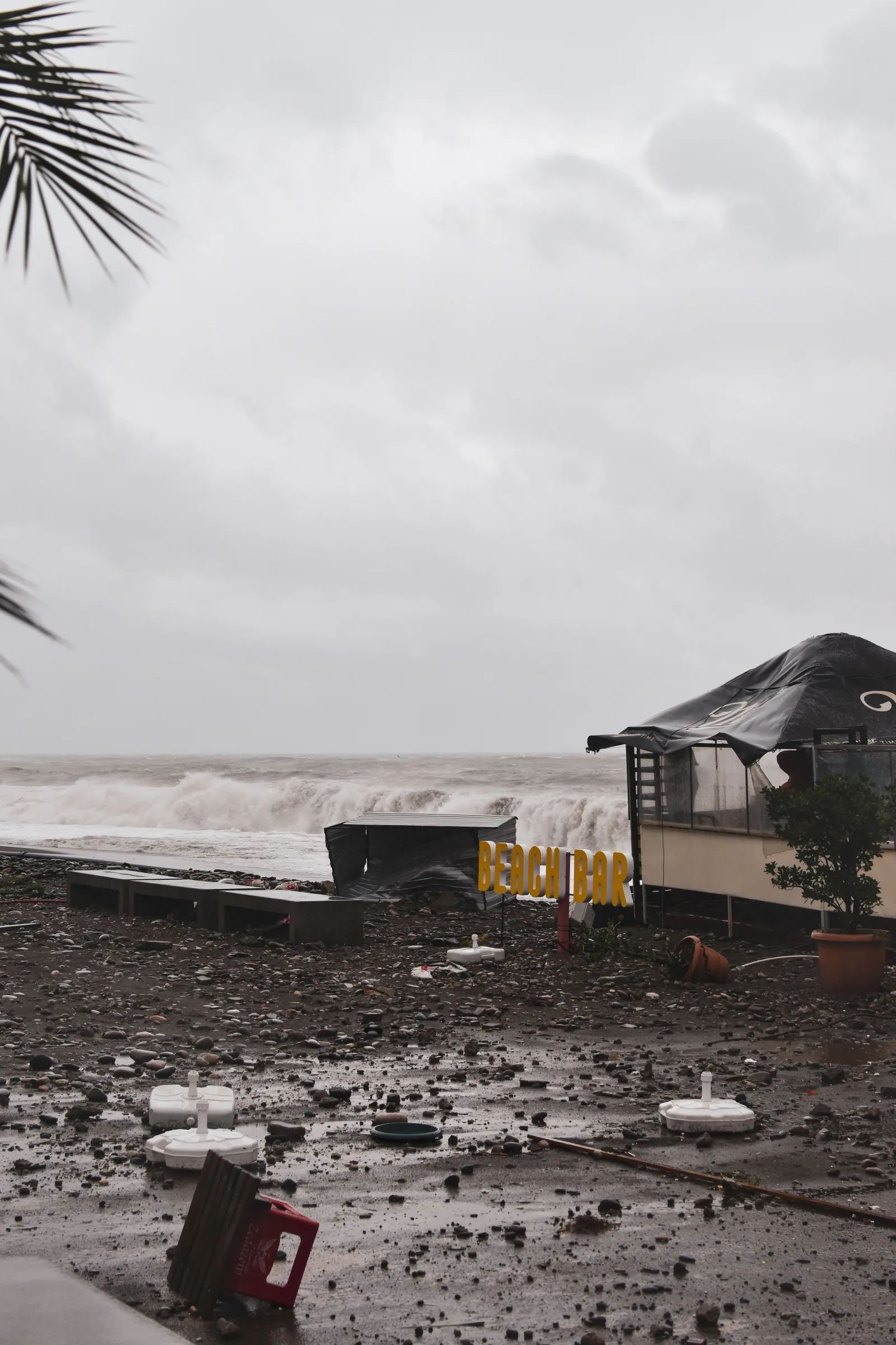 Un bar de playa costero dañado por una tormenta reciente, con olas altas, escombros y cielos nublados.
